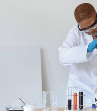 Moça de jaleco, óculos e luvas de plástico mexendo em equipamentos de laboratório - science ambassador
