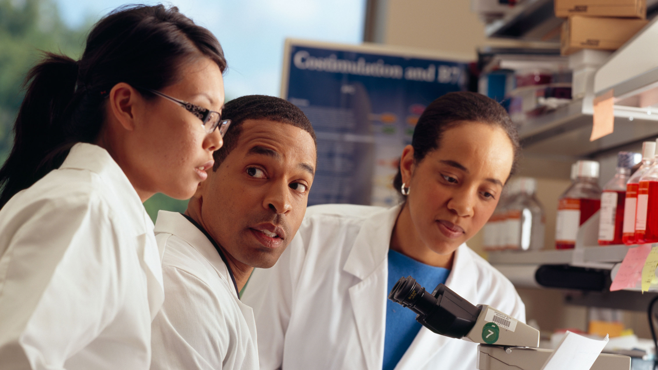Três pessoas de jaleco olhando um microscópio - carreira acadêmica