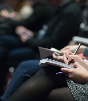 Moça sentada de pernas cruzadas escrevendo num bloquinho em um evento para imprensa - bolsas para jornalistas